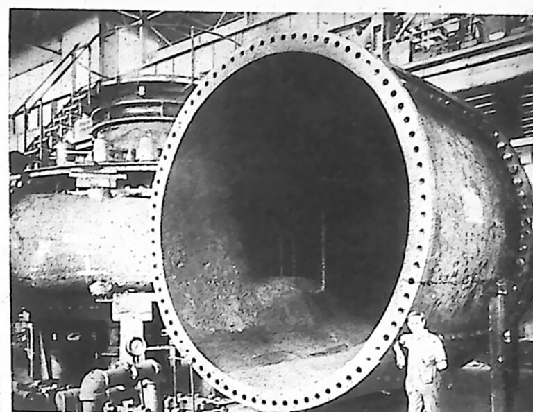 Ogromny cylindryczny kadłub turbiny w hali warsztatowej; obok stoi dorosły mężczyzna, sięgający zaledwie części jej średnicy. Fotografia z 1926 r.