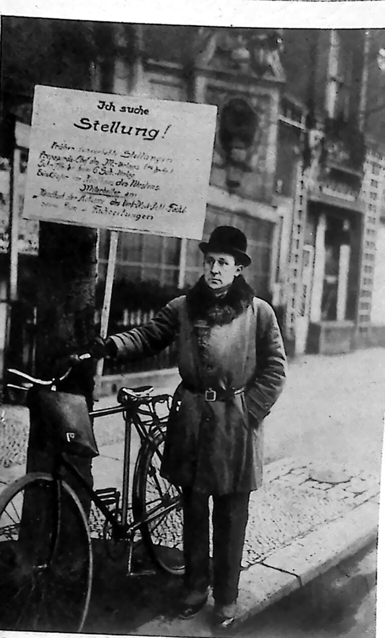Mężczyzna w płaszczu i kapeluszu przy rowerze, z dużą tablicą „Ich suche Stellung!” na ulicy Berlina. Fotografia z 1926 r.