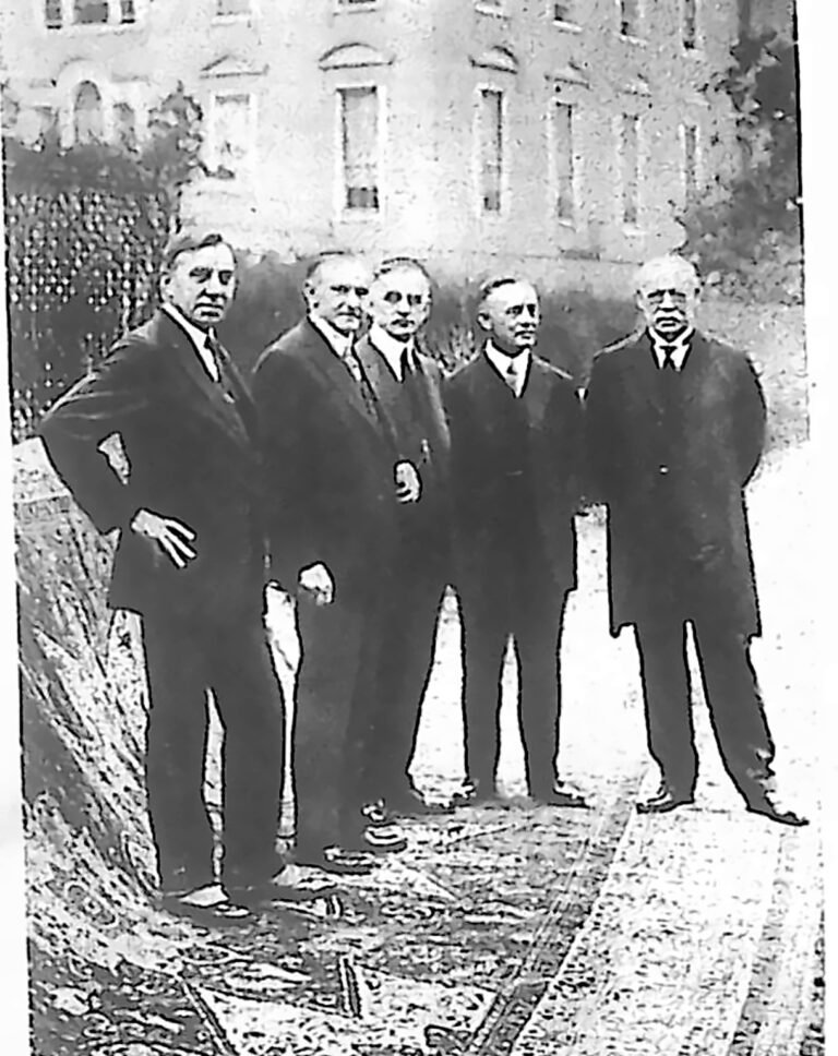 Grupa mężczyzn w garniturach stojąca na rozłożonym ręcznie tkanym dywanie przed budynkiem. Fotografia z 1926 r.