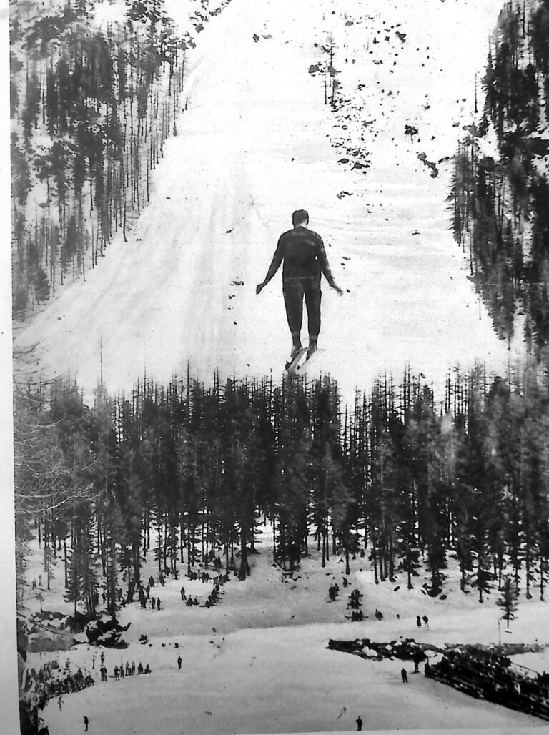 Narciarz w locie nad zaśnieżonym zboczem i lasem świerkowym podczas skoku na odskoczni Bernina w Pontresinie. Fotografia z 1926 r.