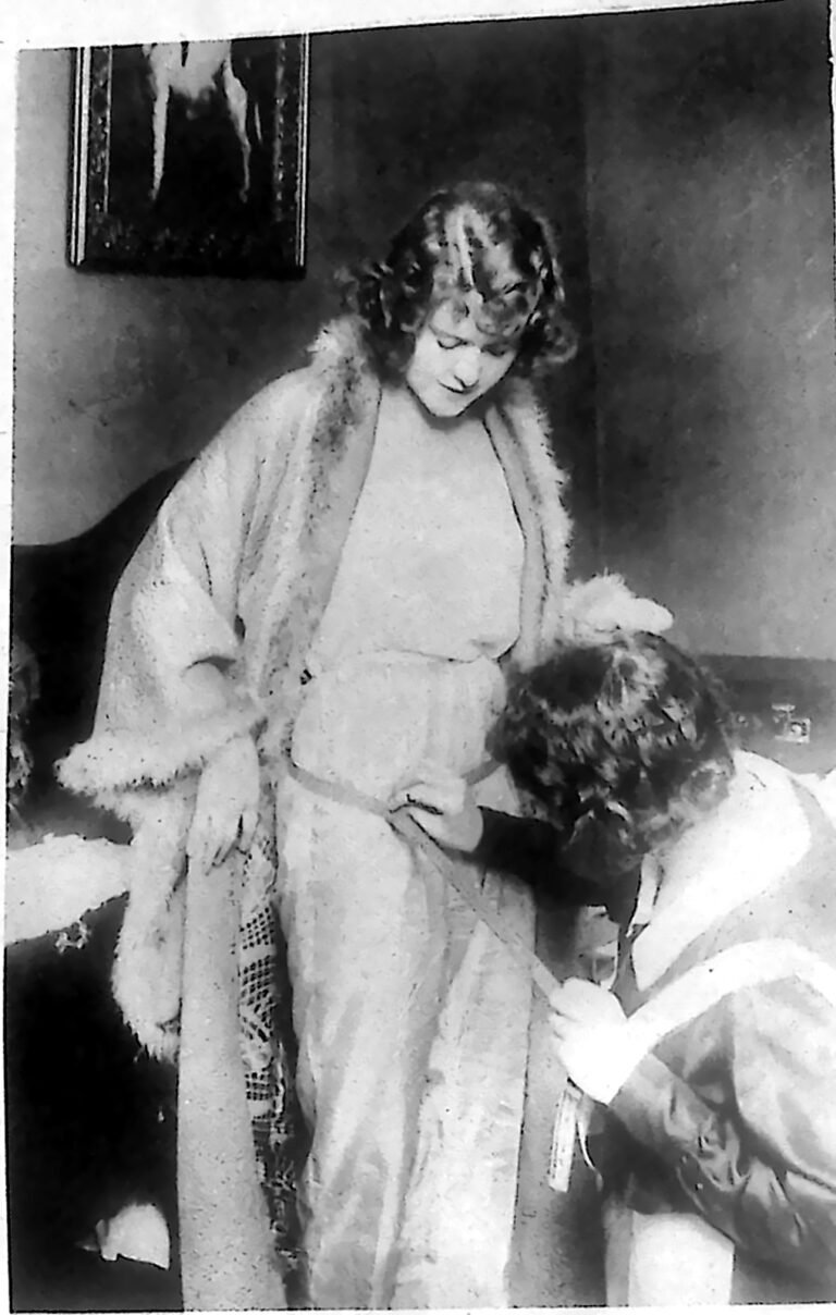 Aktorka w futrze stojąca w pokoju, mierzona w pasie taśmą przez pochyloną pokojówkę. Fotografia z 1926 r.