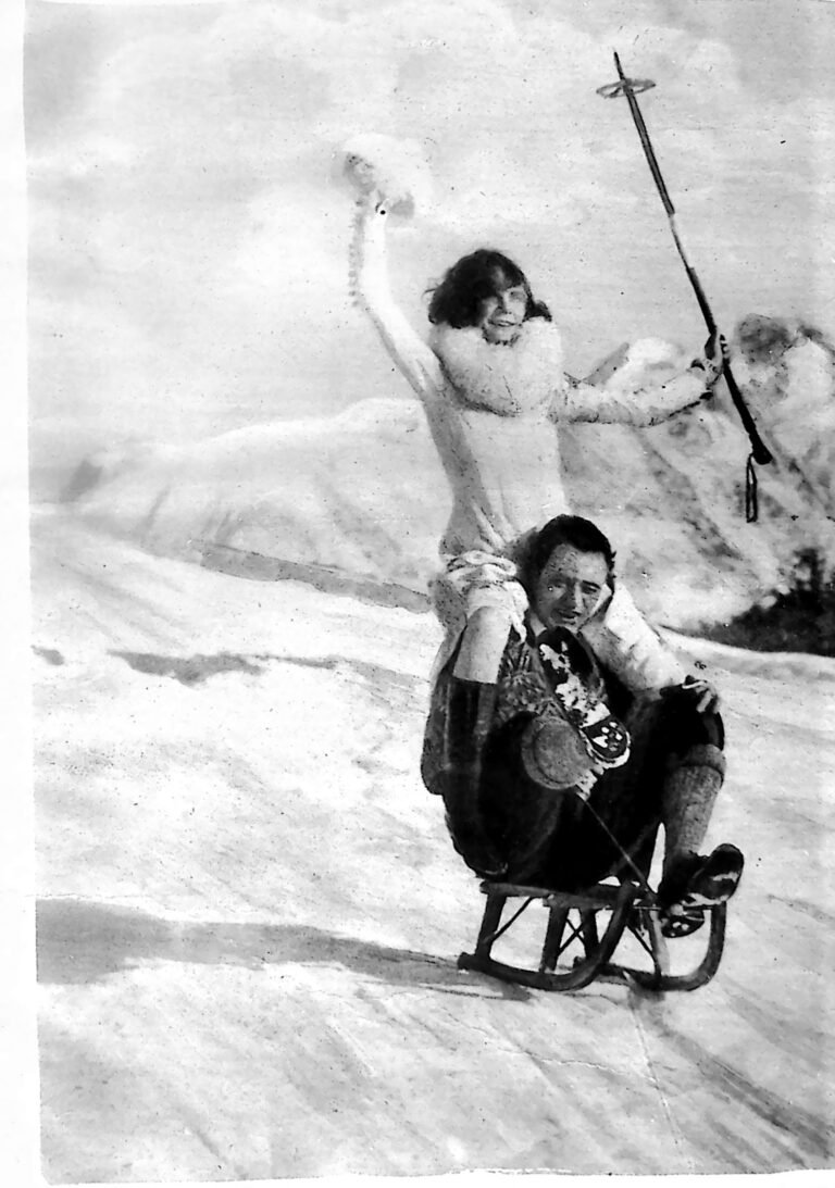 Dwie roześmiane narciarki na saneczkach na zaśnieżonym stoku w Davos, jedna z nich wznosi kijek do góry. Fotografia z 1926 r.
