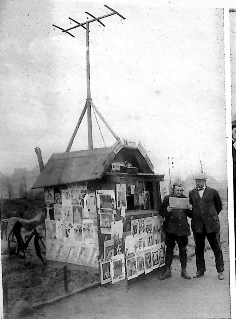 Berliński kiosk z gazetami obwieszony ilustrowanymi pismami, z anteną radiową na dachu; obok dwaj mężczyźni. Fotografia z 1926 r.
