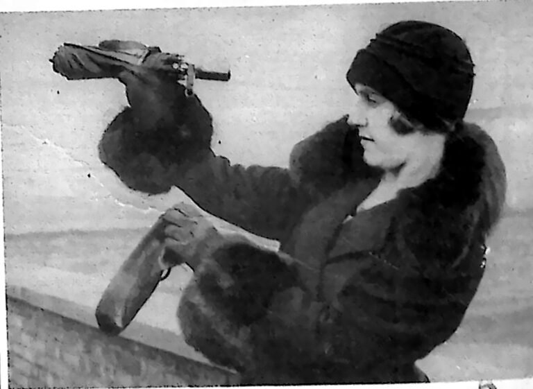 Kobieta w kapeluszu i futrze trzymająca parasolkę z wbudowaną lunetą, przez którą spogląda. Fotografia z 1926 r.