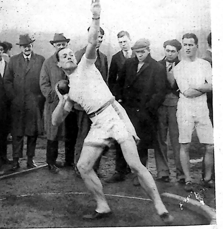 Zawodnik w stroju sportowym w chwili wypchnięcia kuli, otoczony widzami w paltach i kapeluszach. Fotografia z 1926 r.