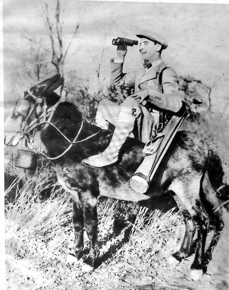 Komik filmowy Johnny Hines w czapce, siedzący na osiołku z lunetą przy oku i torbą kijów golfowych. Fotografia z 1926 r.