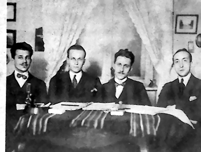 Czterej młodzi mężczyźni w garniturach za stołem prezydialnym, przed nimi udekorowany sztandar. Fotografia z 1926 r.