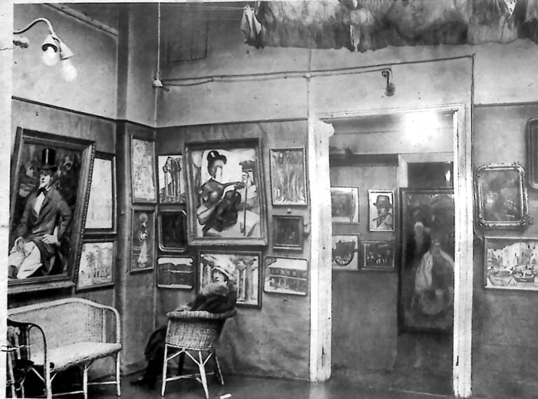 Wnętrze salonu wystawowego z obrazami zawieszonymi gęsto na ścianach i wiklinowymi krzesłami pośrodku. Fotografia z 1926 r.