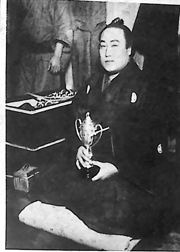 Japoński zapaśnik w ciemnym kimonie, siedzący z pucharem w wyciągniętej dłoni. Fotografia z 1926 r.