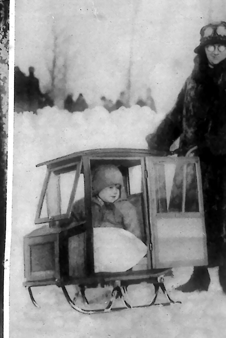 Dziecko siedzące w przeszklonych saniach przypominających karoserię limuzyny, obok dorosły w futrze. Fotografia z 1926 r.