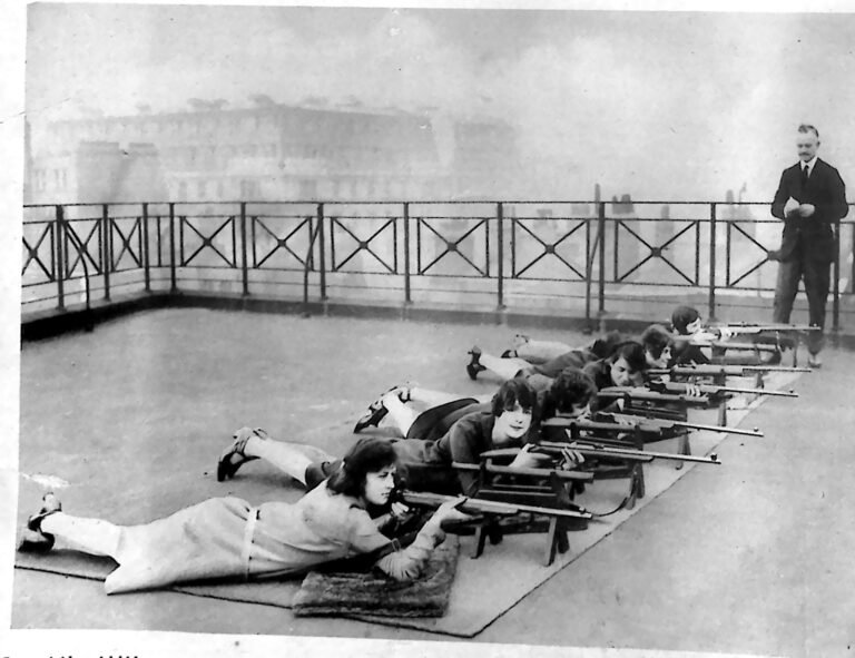 Rząd kobiet leżących na dachu budynku i strzelających z karabinków, obok instruktor w garniturze. Fotografia z 1926 r.