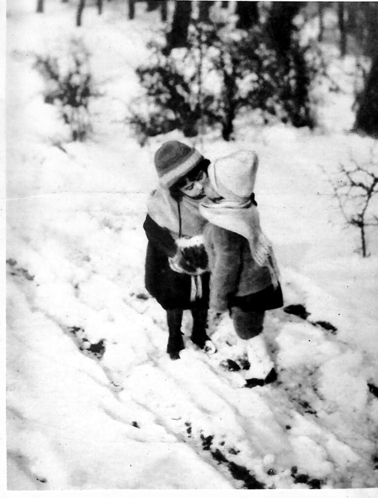 Dwoje małych dzieci w czapkach i szalikach całujących się na zaśnieżonej ścieżce wśród drzew. Fotografia z 1926 r.