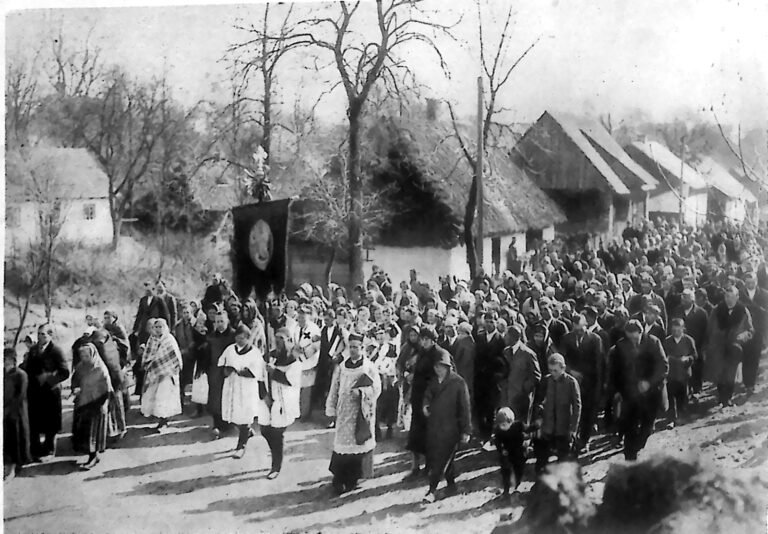 Procesja mieszkańców Bronowic drogą wiejską wśród krytych strzechą chat, na czele duchowni w ornatach. Fotografia z 1926 r.