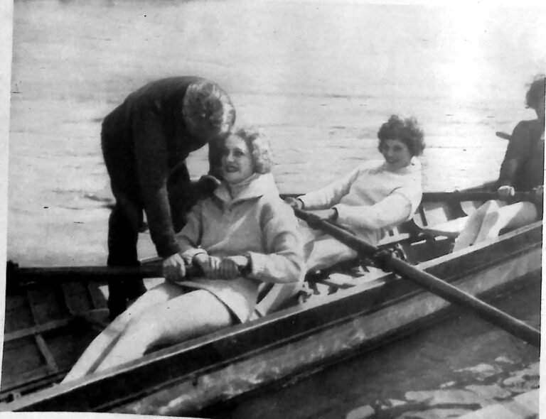 Cztery kobiety w łodzi wioślarskiej na rzece, instruowane przez pochylonego nad nimi trenera. Fotografia z 1926 r.