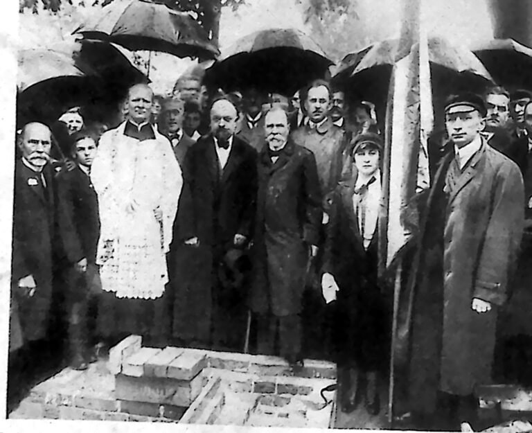 Duchowny i grupa gości pod parasolami przy kamieniu węgielnym na placu budowy w Warszawie. Fotografia z 1926 r.