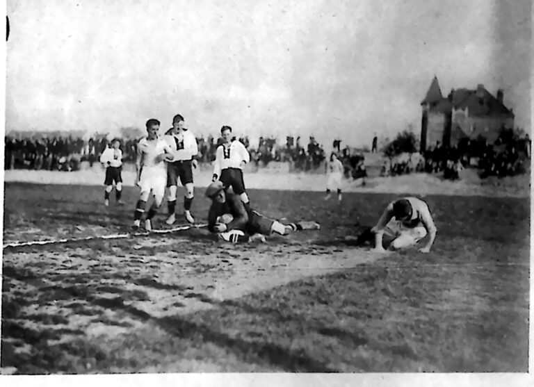 Piłkarze w walce o wysoką piłkę na boisku, w tle tłum widzów i zabudowania. Fotografia z 1926 r.