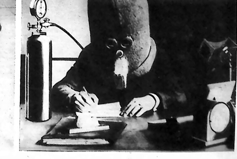 Mężczyzna piszący przy biurku z głową zamkniętą w dużym hełmie izolacyjnym, obok butla z gazem. Fotografia z 1926 r.