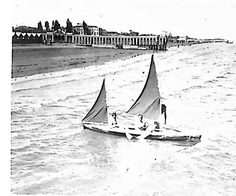 Żaglowa łódka z dwiema osobami na spokojnej wodzie u wybrzeża Lido, w tle zabudowania plażowe. Fotografia z 1926 r.