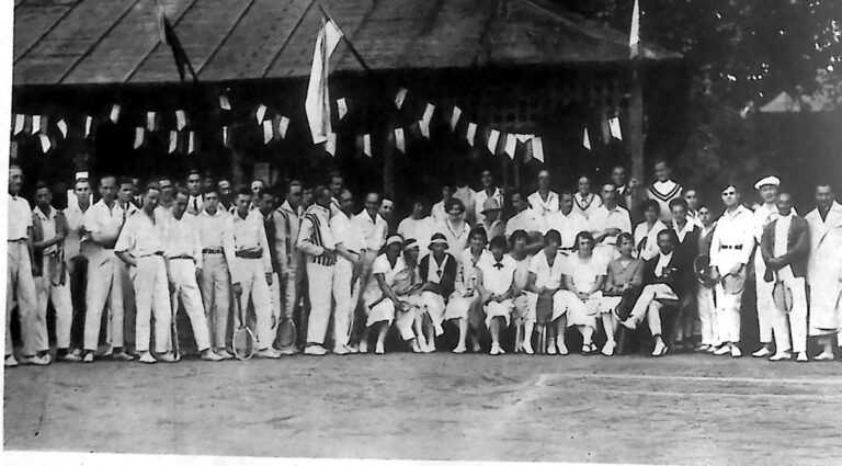 Liczna grupa tenisistów i tenisistek z rakietami przed pawilonem klubowym, przybranym chorągiewkami. Fotografia z 1926 r.